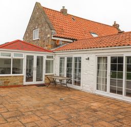A stone house with red tiled roof and glass doors opening to a patio with a wooden table at Jara Bamburgh