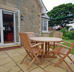 A stone patio with a round wooden table and four wooden chairs outside a stone house with an open door and garden in the background at Stone Cottage in Bamburgh