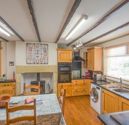 A kitchen with wooden cabinets a dining table a washing machine and a window with blinds at Creel Cottage Amble