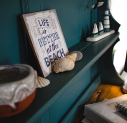 A shelf with a sign saying life is better at the beach seashells a small model lighthouse and boats next to a lamp on a wooden surface at Baysyde in Alnmouth
