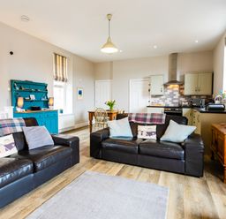 An open plan living room and kitchen with two black leather sofas a wooden dining table blue cabinet and kitchen appliances at Baysyde Alnmouth