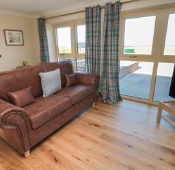 A living room with a brown leather sofa, blue plaid curtains, wooden floor, a TV on a wooden stand and large windows at Nook Cottage in Embleton