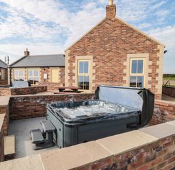 An outdoor area with a hot tub sunken into a brick enclosure and a brick building with windows and a picnic table at Byre Cottage in Embleton