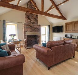 A living area with brown sofas a brick fireplace a dining table with chairs wooden cabinets and a wall mounted television at Smithy Cottage in Embleton