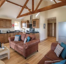 An open plan kitchen and living room with wooden beams on the ceiling brown sofas and wooden cabinets at Smithy Cottage in Embleton