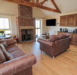 A living room with brown leather sofas a brick fireplace a dining table and wooden kitchen cabinets at Bothy Cottage in Embleton