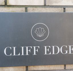 A black rectangular sign with a shell logo and the text Cliff Edge mounted on a brick wall at 33 Cliff Edge in Newquay