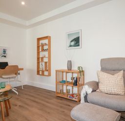 A room with a desk and chair a beige armchair with ottoman and wooden shelves at 33 Cliff Edge in Newquay