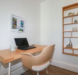 A home office with a laptop on a wooden desk and a wooden chair next to a wall-mounted wooden shelf at 33 Cliff Edge in Newquay
