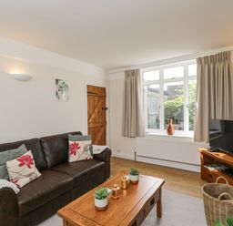 A living room with a brown leather sofa pillows a wooden coffee table with plants a television on a wooden stand and a large window at 2 Church Street in Chipping Norton