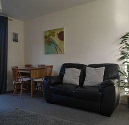 A living room with a black leather sofa and two cushions next to a dining table with wooden chairs and a potted plant at Cliffwalk Cottage in Southwell