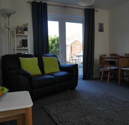 A living room with a black sofa with green cushions a wooden dining table with chairs and a small white side table with fruit at Cliffwalk Cottage in Southwell