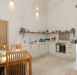 A kitchen with white cabinets a wooden dining table and chairs and shelves with kitchen items at Meadow Syke Barn in Bampton Cumbria
