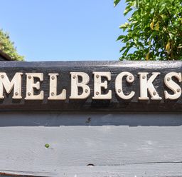 A black wooden sign with white letters spelling Melbecks mounted on a grey wooden fence with greenery and blue sky behind at Melbecks in Abergele