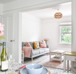 A living room with a gray sofa pink cushions and a coffee table next to a dining table with blue bowls and a wine bottle at River Lym Cottage Lyme Regis