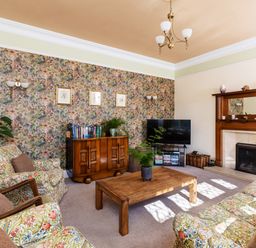A living room with patterned wallpaper a wooden coffee table floral armchairs a flat screen tv and a fireplace at Kail House in Grassington