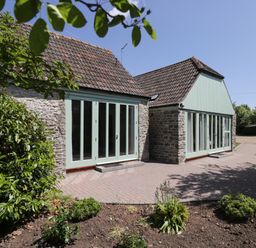 The exterior of a stone building with green framed large windows and doors with a paved pathway and some garden plants at Swallow Barns in Chipping Sodbury