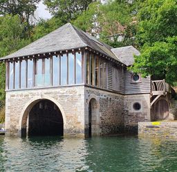 A stone and wood boathouse on a body of water with glass windows and surrounding trees at Oakenhurst in Golant