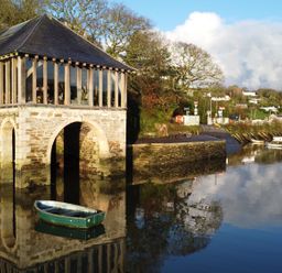 A stone building with arched openings by the water with a small rowboat floating nearby and several boats moored in the background at Oakenhurst in Golant