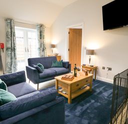 A living room with two blue sofas a wooden coffee table with wine glasses a tv mounted on the wall and a stainless steel fridge at Bracken Cottage in Pocklington