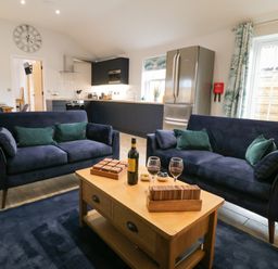 A living room with two navy sofas a wooden coffee table with wine glasses and a bottle and a kitchen with appliances in the background at Bracken Cottage in Pocklington
