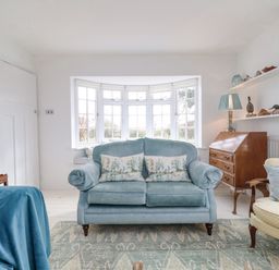A living room with a blue sofa and cushions a wooden secretary desk and shelves with decorative items at Halsey in Walberswick