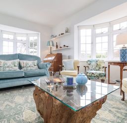 A living room with a blue sofa, armchairs, a glass coffee table, and windows at Halsey in Walberswick