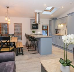 An open plan kitchen and dining area with a wooden dining table and black chairs a kitchen island with bar stools under ceiling lights and skylight a potted orchid on a coffee table at 25 Thirlmere in Troutbeck Bridge