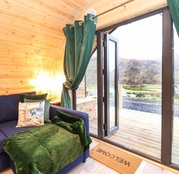 A wooden interior room with a blue sofa and green curtains next to open glass doors opening to an outdoor deck at The Shearer - Crossgate Luxury Glamping in Hartsop near Glenridding