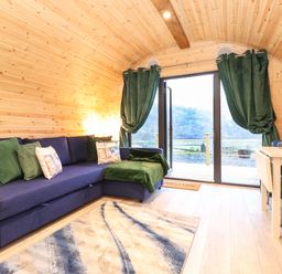 A living area with a blue sectional sofa with green cushions a rug and glass doors with green curtains at The Shearer - Crossgate Luxury Glamping in Hartsop near Glenridding