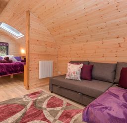 A room with wooden walls a gray sofa with pillows a patterned rug and a bed with purple bedding at Lovies Place - Crossgate Luxury Glamping in Hartsop near Glenridding
