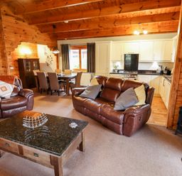 An open living room with brown leather sofas a wooden dining table with chairs a kitchen with white cabinets and a black stove at Avocet Lodge in Tattershall Lakes Country Park