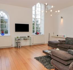 A living room with two arched windows a wall mounted TV and brown sofas at 2 The Reformed Church in Embleton
