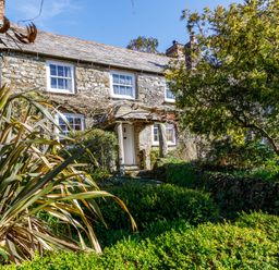 A stone cottage with white framed windows and a garden with bushes and trees at Aarons Cottage in St Teath