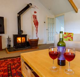 A living room with a wood stove a television a wooden table with two glasses of red wine and colorful cushions on a white sofa at Aarons Cottage in St Teath
