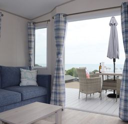 A living room with a blue sofa and wooden coffee table next to a sliding door opening to a deck with wicker chairs and a table with a bottle and glasses at The Retreat in Otterton