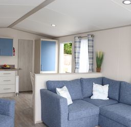 A living room with blue sectional sofa two pillows blue plaid curtains partition wall and kitchen with blue cabinets at The Retreat in Otterton