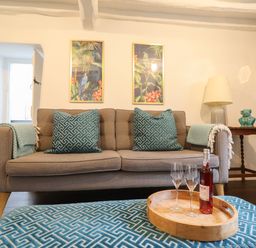 A living room with a brown sofa with teal patterned pillows and blankets a wooden side table with a lamp and decorative item two framed pictures and a wooden tray on a patterned teal ottoman with a wine bottle and two glasses at 3 Ivy Court in Dolgellau