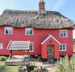 A red cottage with a thatched roof wooden door and a wooden well structure in front at Rhubarb Cottage in Ufford near Woodbridge