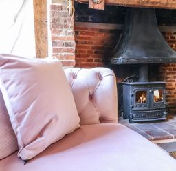 A pink sofa with cushions near a black wood burning stove in a brick fireplace at The Bridewell in Woodbridge