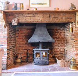 A brick fireplace with a black wood stove and wooden mantel with decorative items at The Bridewell in Woodbridge