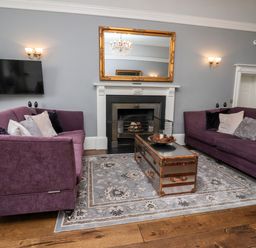 A living room with two purple sofas a fireplace a gold-framed mirror and a chest-style coffee table at The Stables and West Wing Bolton Percy near Tadcaster