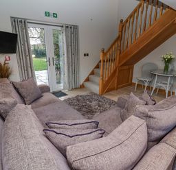 A living room with a large gray sofa a wooden staircase a small round table with chairs and glass doors leading outside at Servant's Quarters Bolton Percy near Tadcaster