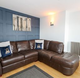 A living room with a brown leather sectional sofa and blue cushions in front of a blue paneled wall with wall art at Pointers View in Weymouth