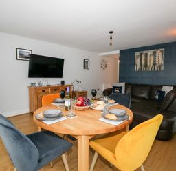 A dining area with a round wooden table set for four and colorful chairs next to a living room with a dark sofa and TV at Pointers View in Weymouth
