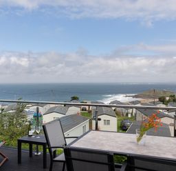 A balcony with a table, chairs, flowers, and a bottle of wine overlooking a caravan park and the sea at Treen in St Ives