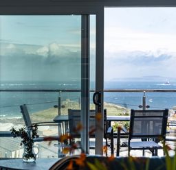 A balcony with chairs and a table overlooking the sea through glass doors at Treen in St Ives