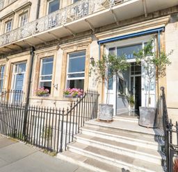 A building facade with stairs leading to a doorway flanked by two potted plants and window boxes with flowers at The Esplanade in Weymouth