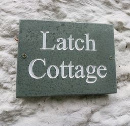 A green plaque with the words Latch Cottage on a white textured wall at Latch Cottage in Keswick
