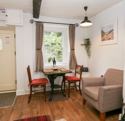 A small dining area with two wooden chairs with red cushions around a wooden table near a window with curtains and an armchair in a room at Latch Cottage in Keswick